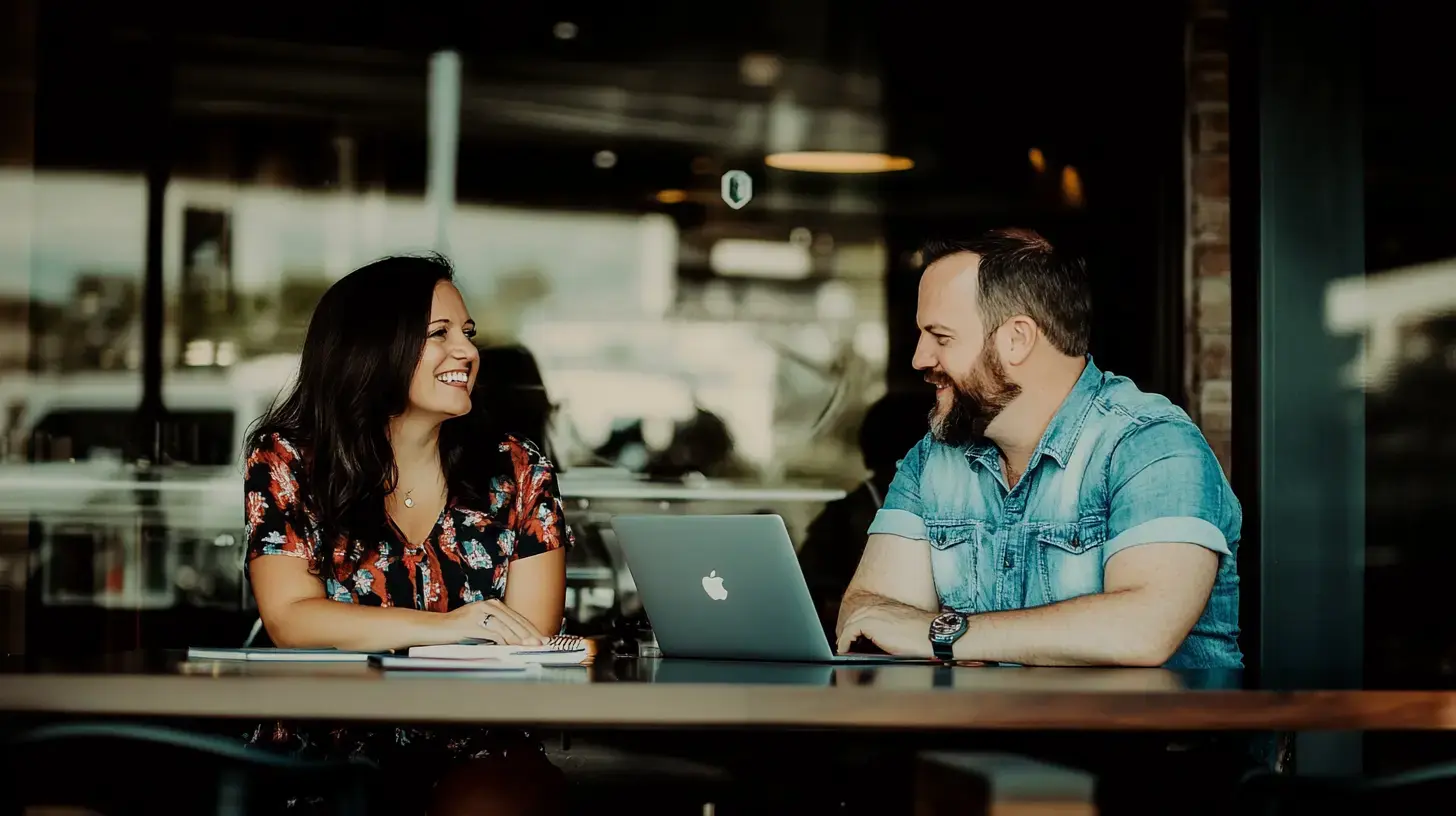 Two people converse at a coffee shop, smiling and listening, with a laptop and notebooks on the table.