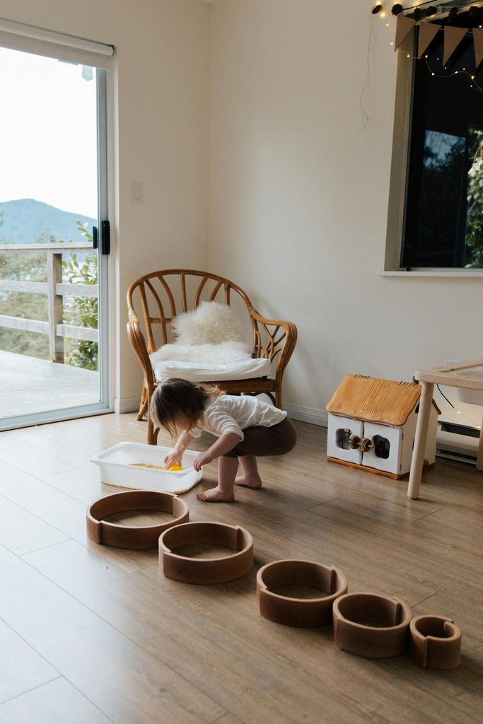Full length back view of faceless toddler standing barefoot on floor playing with round wooden shapes of different size and using white container while developing fine motor skills at home