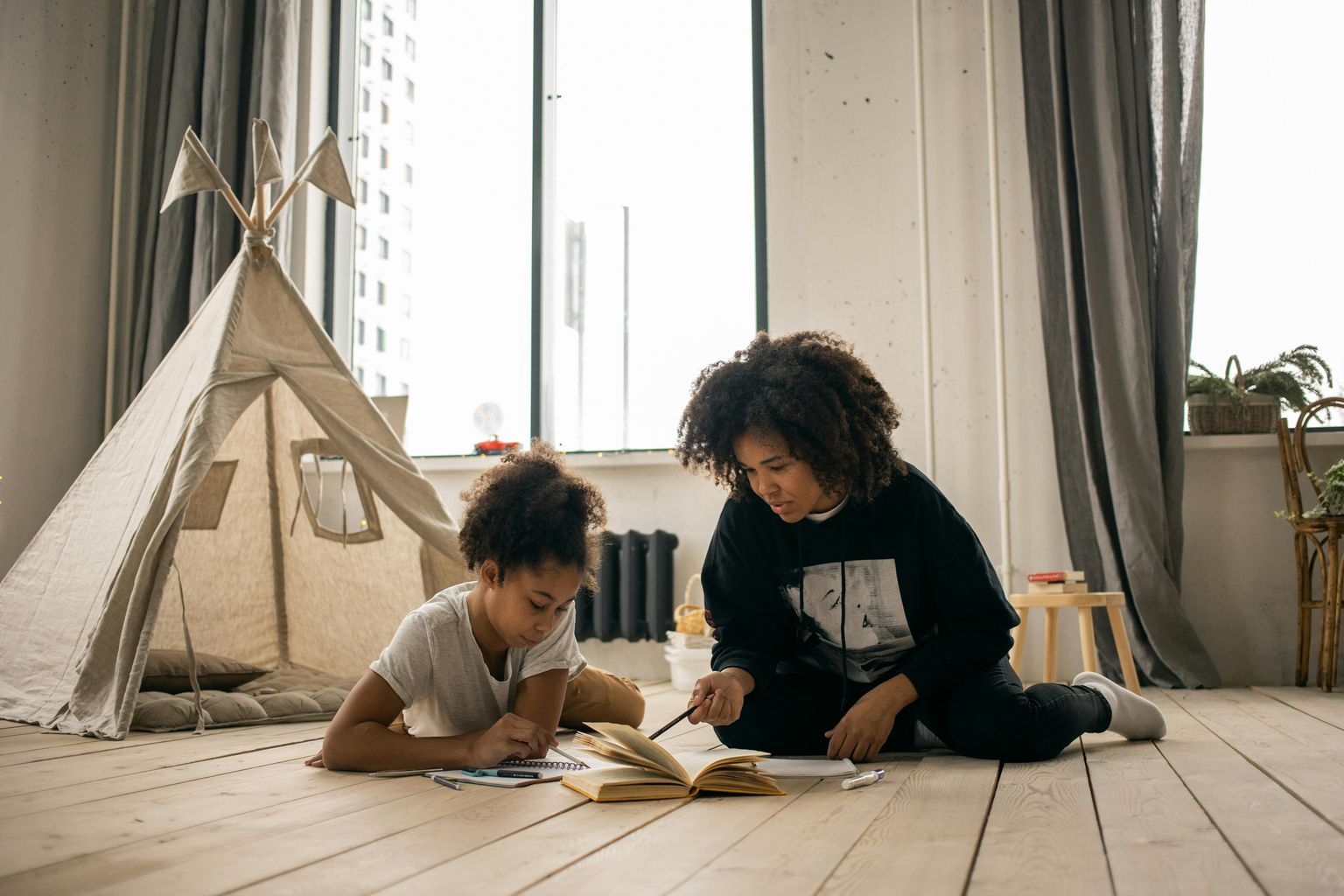 African American woman with Afro hairstyle doing homework with daughter lying on floor in kid bedroom